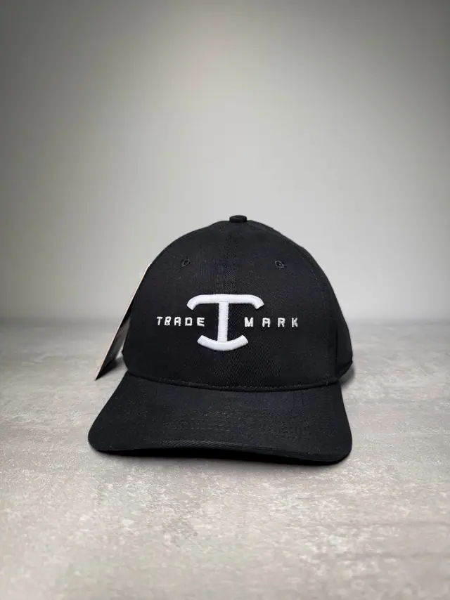 Boné preto de aba curva com o bordado branco TRADE MARK atravessado ao centro pelo monograma T da TBull, ainda com a tag de papel pendurada na lateral da aba.
