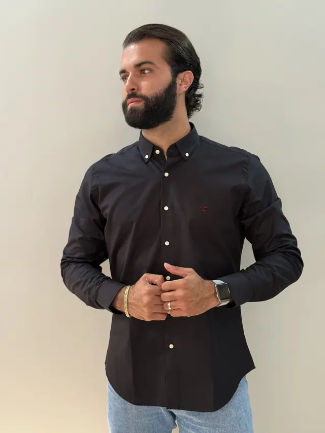 Modelo veste a camisa Preto OX TP: oxford preta de manga longa, colarinho button-down e monograma bordado em vermelho no peito.