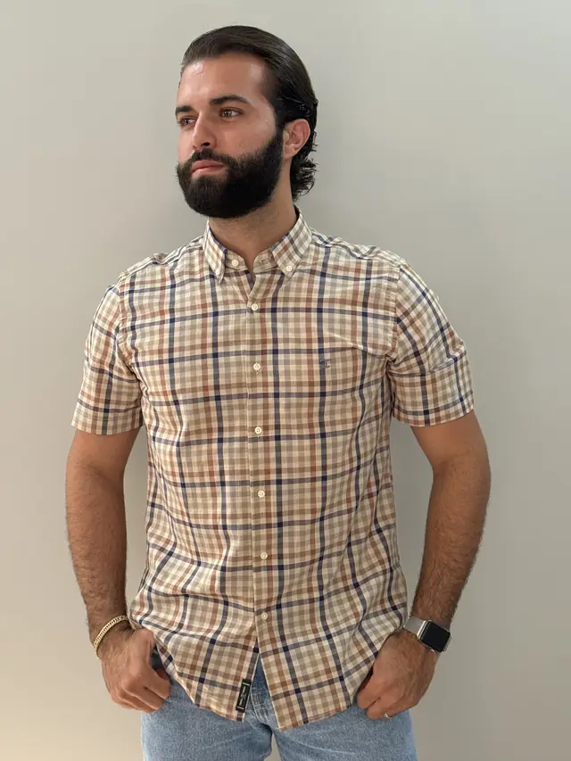 Modelo veste a camisa Cacau: xadrez de fundo bege riscado em cacau e marinho, manga curta e bolso frontal.