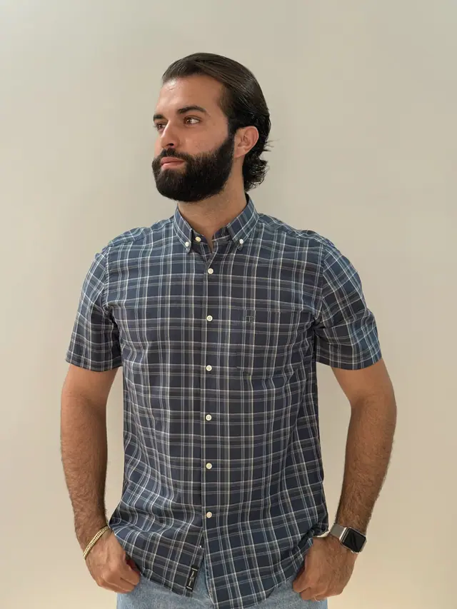 Modelo veste a camisa Xadrez 1226: xadrez azul-marinho riscado em creme, manga curta e bolso frontal.