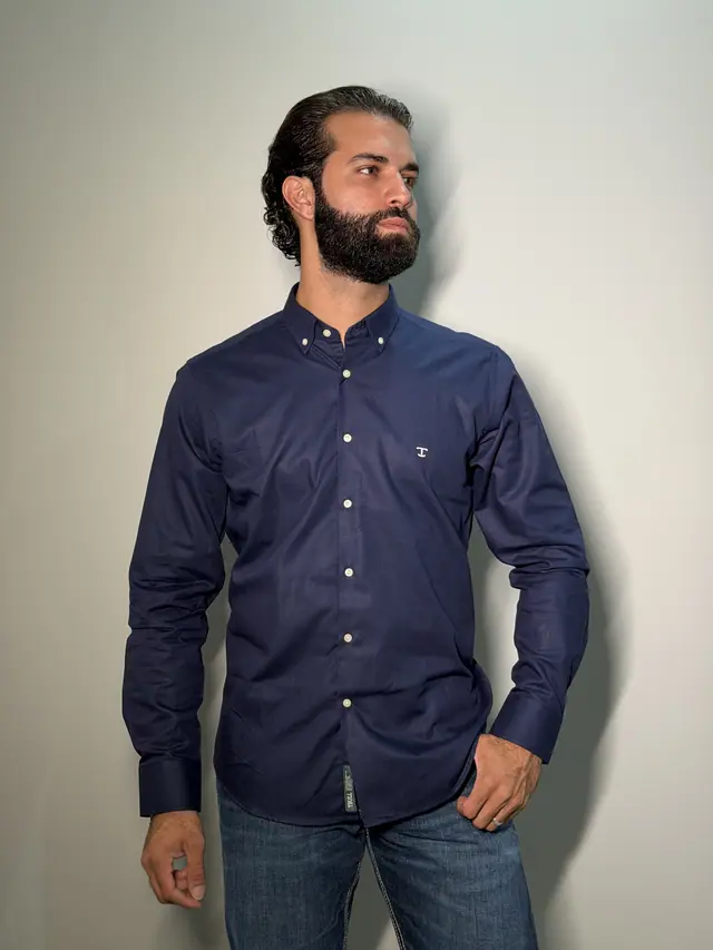 Homem de camisa social de manga longa azul-marinho, gola button-down com botões claros, de frente e com o rosto virado de lado, contra parede clara.