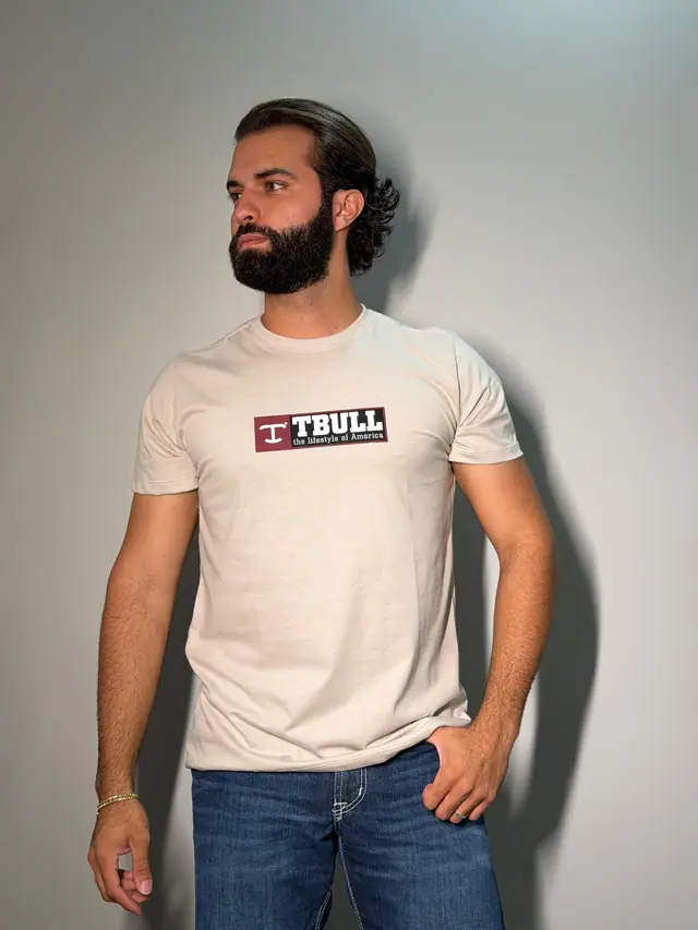 Homem de barba veste camiseta TBull off white de malha, com etiqueta vinho estampada no peito, e calça jeans, em foto de estúdio.