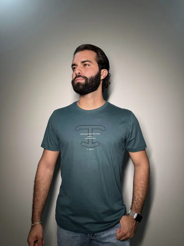 Modelo veste a camiseta Desert: malha verde petróleo com o monograma T em contorno no centro do peito.